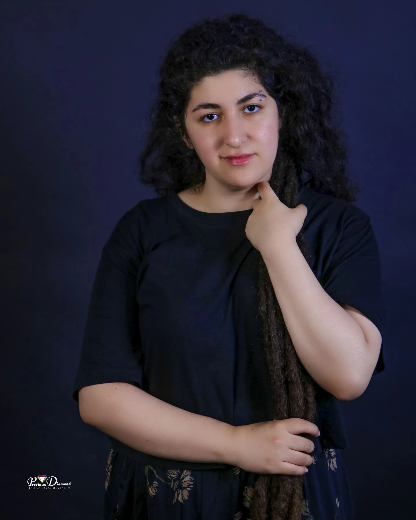 Portrait on a navy backdrop, one hand resting near the chin, long locs over one shoulder