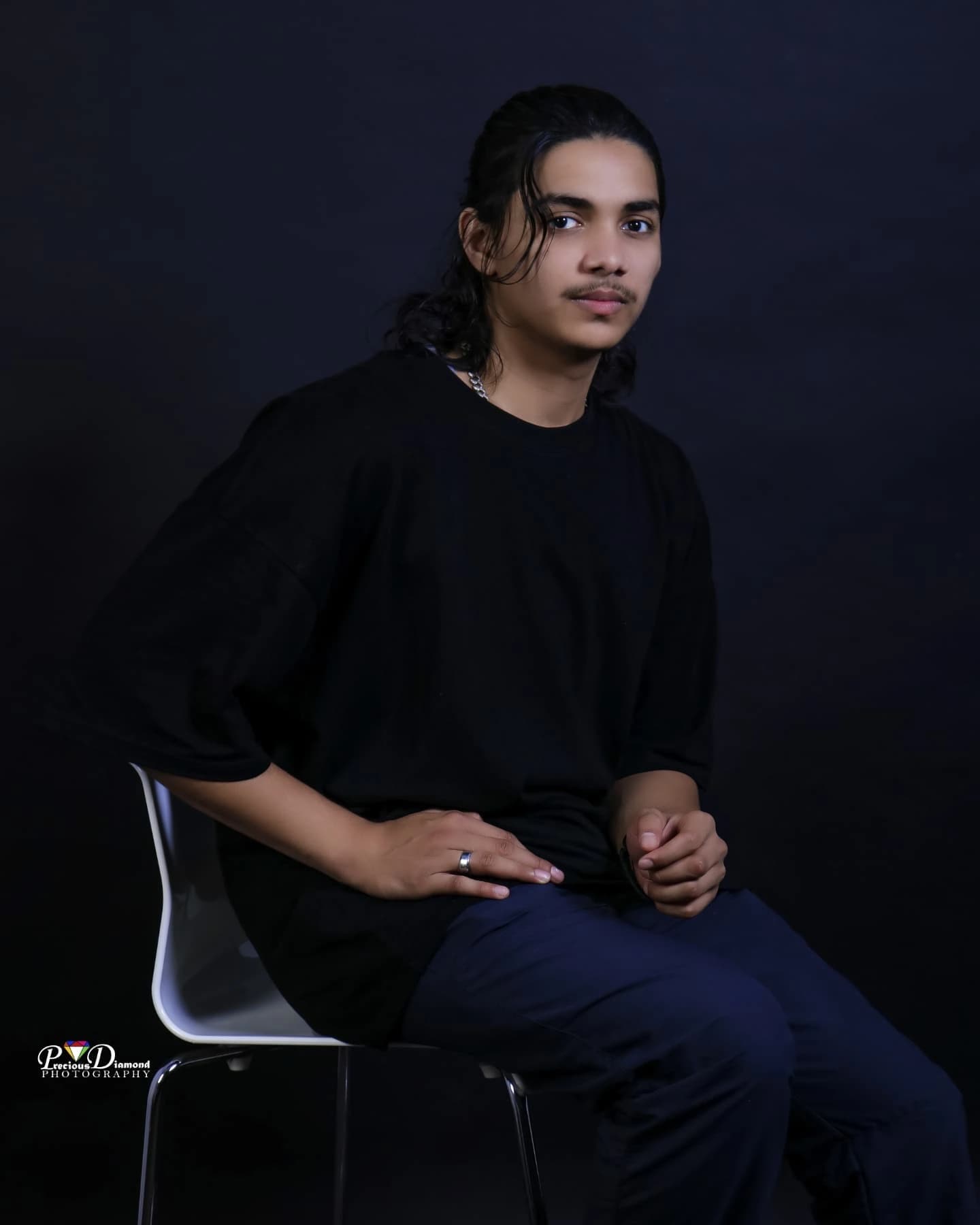 Seated portrait on a navy backdrop, leaning forward on a white chair with hands clasped
