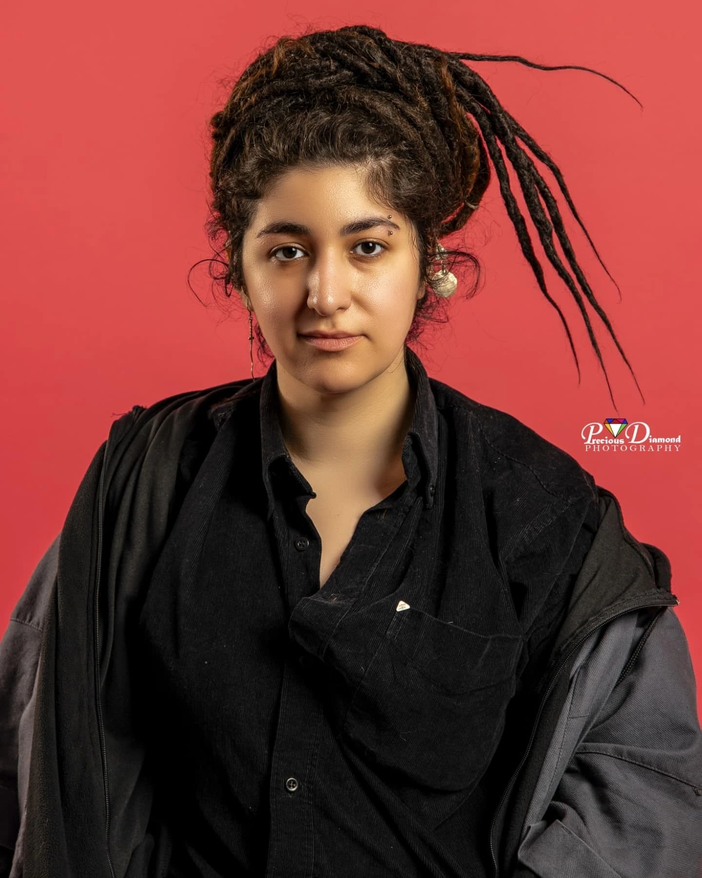 Studio portrait on red with locs piled high and swept to one side, in a black shirt and coat